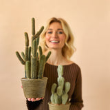 Een glimlachende vrouw in een bruine jurk houdt twee Kunst Cactus Cereus 35 cm planten in pot, een grote en een kleine, tegen een effen beige achtergrond.