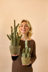 Een glimlachende vrouw in een bruine jurk houdt twee Kunst Cactus Cereus 35 cm planten in pot, een grote en een kleine, tegen een effen beige achtergrond.