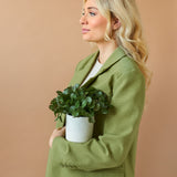 Een vrouw met lang blond haar in een lichtgroene blazer houdt de "Kunst vetplant 22 cm in sierpot" tegen haar borst en staat zijdelings voor een effen beige achtergrond.