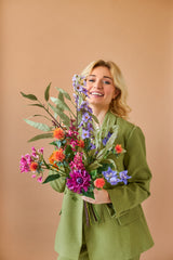 Een lachende blonde persoon in een groen pak houdt het levendige Zijden kunstbloemen boeket Colourful Svenski tegen een effen, lichtbruine achtergrond.