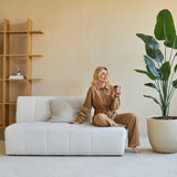 Een vrouw in een bruine outfit zit op een witte bank, glimlachend met een kopje in haar hand. Naast haar staat de Kunstplant Strelitzia 180 cm real touch, die weelderig groen brengt in de moderne, beige getinte woonkamer met minimale inrichting.