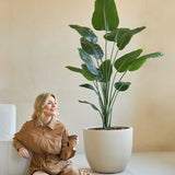 Een vrouw in een bruine jumpsuit zit glimlachend op de grond met een koffiekopje naast zich, naast de Kunstplant Strelitzia 180 cm real touch in een minimalistische kamer met beige muren.