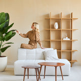 Een vrouw in een bruine jumpsuit zit op de rugleuning van een witte bank en raakt het blad aan van een Kunstplant Strelitzia Real Touch Deluxe 180 cm in een minimalistische woonkamer met houten tafels en planken.