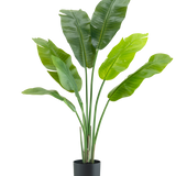 De Kunstplant Strelitzia Nicolai Deluxe 120 cm is een luxe kamerplant met grote, brede bladeren in een eenvoudige zwarte pot, stijlvol tegen een witte achtergrond.