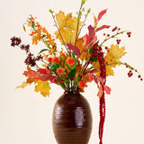 In een bruine vaas staat het boeket ‘Rustic Glow’ van Zijden kunstbloemen, met gele, oranje en rode bladeren, rode bessen, oranje bloemen en lange, hangende rode amaranthus tegen een neutrale achtergrond.