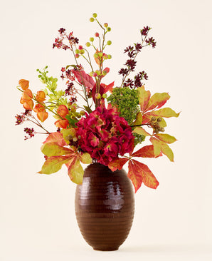 In een bruine, geribbelde vaas staat het boeket „Zijden kunstbloemen Warm Amber”, met levendige rode, oranje en groene bloemen en bladeren aan lange stelen, tegen een effen lichte achtergrond.