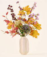 Het boeket ‘Wild Amethyst’ van Zijden kunstbloemen, met herfstbladeren, paarse en witte bloemen en groene hortensia’s, staat in een witte keramische vaas tegen een effen, lichte achtergrond.