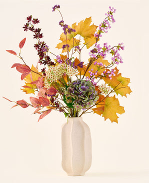 Het boeket ‘Wild Amethyst’ van Zijden kunstbloemen, met herfstbladeren, paarse en witte bloemen en groene hortensia’s, staat in een witte keramische vaas tegen een effen, lichte achtergrond.