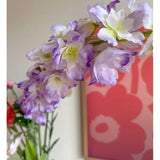 Close-up van de Kunstbloem Delphinium 105 cm paars, geschikt in een vaas, met op de achtergrond een roze en rood bloemenkunstwerk aan de muur.