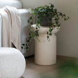 Een Kunst Klimop Deluxe Groen 90 cm staat in een beige vaas op een houten vloer naast een lichtgrijze bank waarop een crèmekleurige plaid ligt en een witte poef met structuur.