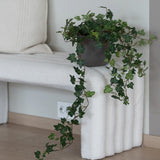De Kunst Klimop Deluxe Groen 90 cm, met zijn weelderige hangende ranken, staat op een wit bankje met structuur naast een crèmekleurig kussen in een minimalistische kamer met lichte houten vloeren.