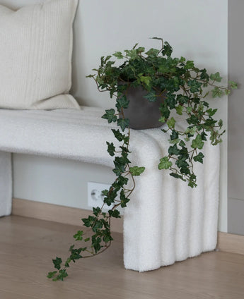 De Kunst Klimop Deluxe Groen 90 cm, met zijn weelderige hangende ranken, staat op een wit bankje met structuur naast een crèmekleurig kussen in een minimalistische kamer met lichte houten vloeren.