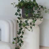 Een Kunst Klimop Deluxe Groen, een hangende klimop van 90 cm in een grijze pot, staat op een modern, wit, sculpturaal voetstuk naast een opgevouwen witte deken en een wit kussen met structuur.