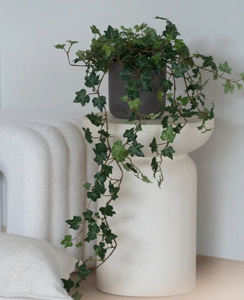 Een Kunst Klimop Deluxe Groen, een hangende klimop van 90 cm in een grijze pot, staat op een modern, wit, sculpturaal voetstuk naast een opgevouwen witte deken en een wit kussen met structuur.