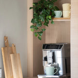 Op het aanrecht staat een zilveren koffiezetapparaat met een groene mok. Daarboven, op een houten plank, staan pastelkleurige kopjes opgestapeld en hangt een ‘Kunst Hangende goudrank 60 cm UV’. Vlak daarnaast staan houten snijplanken tegen de muur geleund.