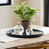 Een Kunst Goudrank groen-wit in pot UV 35 cm staat samen met twee glazen olielampen op een rond spiegelplateau op een houten tafel, verlicht door natuurlijk licht dat door een raam in de buurt naar binnen valt in deze moderne, minimalistische omgeving.