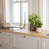 Een lichte keuken met houten werkbladen, witte kasten, twee gouden kandelaars, een witte pan op het fornuis en een groot raam dat natuurlijk licht binnenlaat. De ruimte wordt geaccentueerd door de Kunst Goudrank groen-wit in pot UV 35 cm.