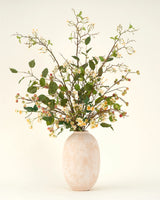 Het Zijden kunstbloemen boeket Wild Apple Blossom, gerangschikt in een hoge vaas van beige keramiek, heeft lange takken met groene bladeren, kleine gele en witte bloemen en trossen ronde bessen tegen een lichte achtergrond.