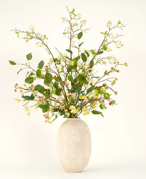 Een hoge vaas van beige keramiek bevat het Zijden kunstbloemen boeket Wild Apple Blossom XL, met groene bladertakken, kleine gele bloemen en roze bessen tegen een lichte achtergrond.