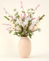 Een vaas met beige structuur bevat het Zijden kunstbloemen boeket Lots of Love in roze, wit en lichtblauw met groene bladeren tegen een effen lichte achtergrond.