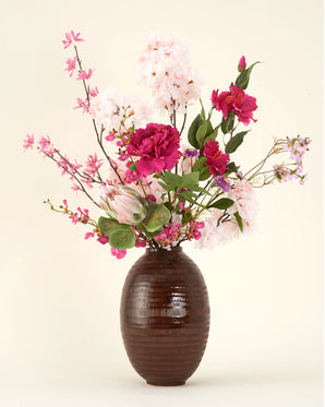 Een bruine geribbelde vaas bevat het Zijden kunstbloemen boeket Pink Reverie, met roze en witte bloemen zoals pioenrozen en kersenbloesems plus groene bladeren, allemaal tegen een effen crèmekleurige achtergrond.