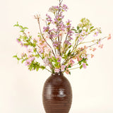 Een bruine vaas van keramiek toont het Zijden kunstbloemen boeket Lilac, een hoog en elegant zijden boeket met roze, paarse en lila bloemen met groene bladeren en dunne takken tegen een effen lichte achtergrond.