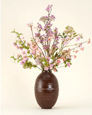 Een donkerbruine vaas van keramiek met textuur bevat het Zijden kunstbloemen boeket Lilac, met roze en lila bloemen met groene bladeren tegen een lichte achtergrond.