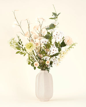 Een witte vaas van keramiek toont het Zijden kunstbloemen boeket Claire de Lune, met zachtgetinte bloemen en weelderig groen gebladerte elegant gerangschikt tegen een neutrale achtergrond.