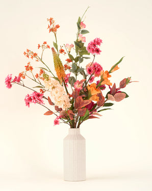 Een witte vaas van keramiek bevat het Zijden kunstbloemen boeket Wild Peach, met levendige zomerbloemen in roze, oranje, geel en crème met groene bladeren, tegen een effen lichte achtergrond.