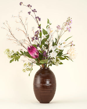 Het Zijden kunstbloemen boeket Serene Bloomfield, met een grote roze protea, witte rozen, paarse bloemen en groen gebladerte in een bruine geribbelde vaas, staat tegen een effen beige achtergrond.