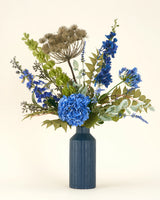 Het Zijden kunstbloemen boeket Mystic Blue Garden, met blauwe hortensia's, riddersporen, groen blad en gedroogde zaaddozen, staat in een hoge, geribbelde donkerblauwe vaas tegen een lichte achtergrond.