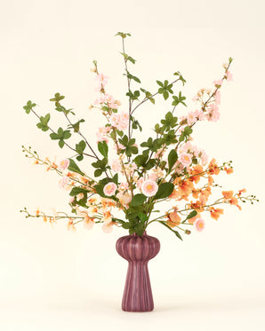 Een mauve vaas toont het Zijden kunstbloemen boeket Blossom Harmony, met roze en oranje bloemen met groen gebladerte. De zijden bloemen zien er delicaat uit, met enkele sierlijke takken tegen een beige achtergrond.