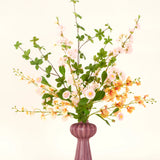 Een Zijden kunstbloemen boeket Blossom Harmony is gearrangeerd in een mauve vaas met roze en perzik bloemen en groene bladeren, tegen een eenvoudige lichte achtergrond voor een elegante display.
