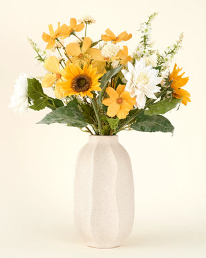 Een crèmekleurige vaas van keramiek toont het Zijden kunstbloemen boeket Sunny Whisper-gele, witte en oranje kunstbloemen met groene bladeren tegen een lichte achtergrond.