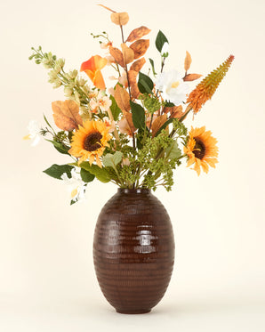Een vaas met bruine structuur toont het Zijden kunstbloemen boeket Amber Light, met zonnebloemen, witte margrieten, oranje lelies en gemengd groen en bruin gebladerte, tegen een eenvoudige lichte achtergrond.