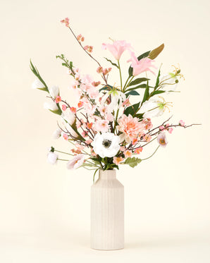 Het Zijden kunstbloemen boeket Pink & Pearl staat in een beige vaas van keramiek, met delicate roze, witte en perzikkleurige bloemen met groen gebladerte tegen een licht crèmekleurige achtergrond.
