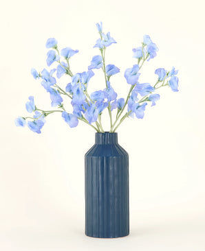 Een blauw geribbelde vaas van keramiek toont het Zijden kunstbloemen boeket Erwt blauw, met zijn delicate lichtblauwe kunstbloemen tegen een effen, lichte achtergrond.