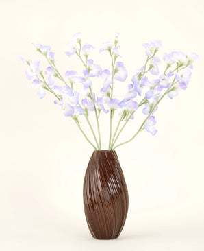 Een bruine, geribbelde vaas bevat het Zijden kunstbloemen boeket Erwt lavendel, met delicate bloemblaadjes en groene stelen tegen een effen gebroken witte achtergrond voor een subtiele cottage sfeer.
