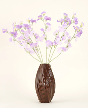 Een bruine vaas met textuur bevat het Zijden kunstbloemen boeket Erwt paars-lichtpaarse zijden bloemen met groenachtige stelen tegen een effen, lichte achtergrond.
