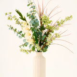 In een beige vaas staat het boeket „Wild Ivory“ van Zijden kunstbloemen, met groene bladeren, eucalyptus, witte bloemen, kleine groene bloesems en lange, dunne bruine grassen tegen een lichte achtergrond.