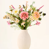 In een witte keramische vaas staat het boeket ‘Backyard Blossom’ van Zijden kunstbloemen, bestaande uit een prachtig arrangement van roze rozen, witte madeliefjes en kleine gele bloemetjes, vervaardigd uit zijden bloemen, aangevuld met weelderig groen blad tegen een effen lichte achtergrond.