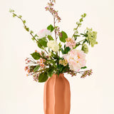 In een perzikkleurige keramische vaas staat een boeket van roze en witte Zijden kunstbloemen, het boeket „Dreamy Meadow”, met groene bladeren en stelen, tegen een effen, lichte achtergrond.
