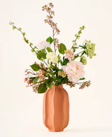 In een perzikkleurige keramische vaas staat een boeket van roze en witte Zijden kunstbloemen, het boeket „Dreamy Meadow”, met groene bladeren en stelen, tegen een effen, lichte achtergrond.