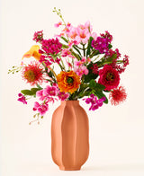 In een perzikkleurige vaas staat het boeket „Tropical Sunset“ van Zijden kunstbloemen, bestaande uit een levendige mix van roze, oranje, gele en rode bloemen tegen een effen, lichte achtergrond.