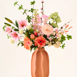 In een terracotta vaas staat het kleurrijke boeket ‘Yasmine’s Choice’ van zijden kunstbloemen, bestaande uit roze lelies, perzikkleurige rozen en groen blad, vervaardigd uit hoogwaardige zijden bloemen van Easyplants, allemaal prachtig gearrangeerd tegen een lichte achtergrond.