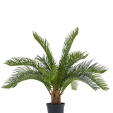 De Kunstpalm Baby Cycus 45 cm heeft lange groene bladeren en een dikke stam in een zwarte pot. Deze onderhoudsvrije kunstplant is een stijlvolle keuze voor elk interieur, vooral tegen een witte achtergrond.
