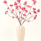 De Zijden kunstbloemen boeket Kersenbloesem fuchsia 114 cm voegt een levendige knal van kleur toe aan je interieur met zijn opvallende takken en fuchsia bloesems, prachtig weergegeven in een beige keramische vaas tegen een effen gebroken witte achtergrond.