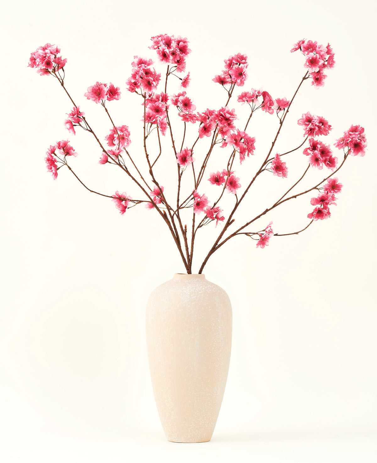 De Zijden kunstbloemen boeket Kersenbloesem fuchsia 114 cm voegt een levendige knal van kleur toe aan je interieur met zijn opvallende takken en fuchsia bloesems, prachtig weergegeven in een beige keramische vaas tegen een effen gebroken witte achtergrond.