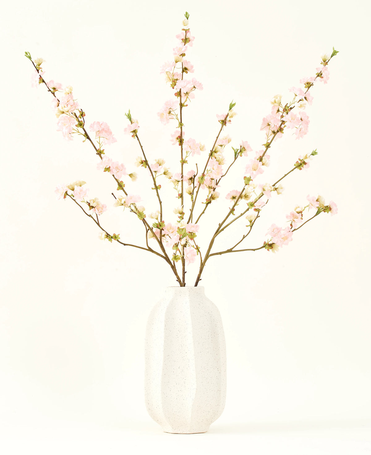 Het Zijden kunstbloemen boeket Kersenbloesem lichtroze 94 cm heeft delicate roze kunstmatige kersenbloesems in een witte keramieken vaas, die elegantie toevoegt tegen een effen lichte achtergrond.