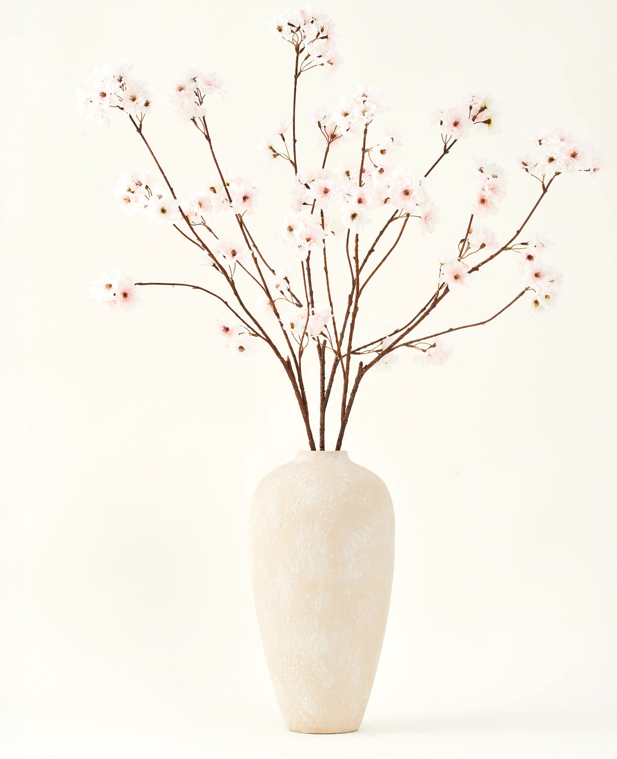 De Zijden kunstbloemen boeket Kersenbloesem blush 114 cm heeft hoge, delicate takken in een eenvoudige beige vaas, die een zachte, elegante touch toevoegt aan je interieur tegen een effen, lichte achtergrond.